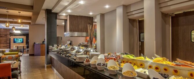 Hotel Lucerna Ciudad Juarez – barra de desayuno tipo buffet con frutas, cereales y platos calientes junto a cocina abierta.