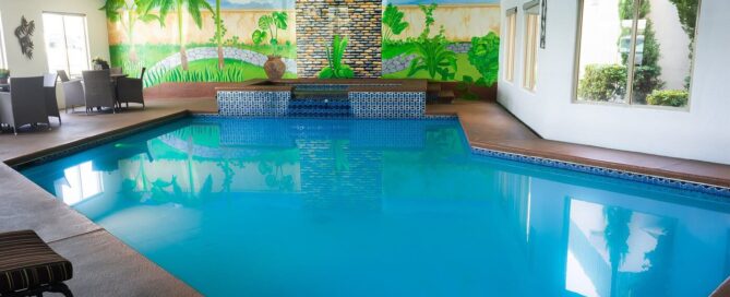 Alberca techada con mural tropical, jacuzzi integrado y mesas con sillas junto a ventanales en María Bonita Business Hotel & Suites.