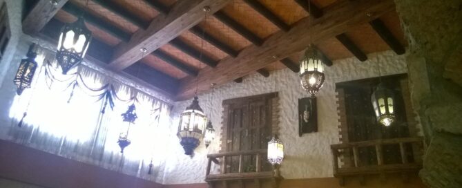 Detalle interior con vigas de madera y lámparas colgantes de hierro forjado, ventanas con cortinas y balconcillos decorativos.