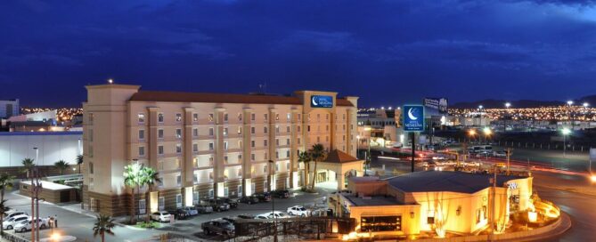 Fachada del Hotel Mesaluna de noche en Ciudad Juárez, iluminación cálida, amplio estacionamiento y letreros visibles junto a la zona del Consulado.