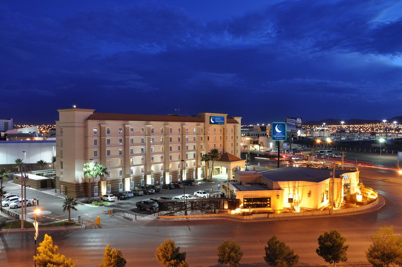 Fachada del Hotel Mesaluna de noche en Ciudad Juárez, iluminación cálida, amplio estacionamiento y letreros visibles junto a la zona del Consulado.