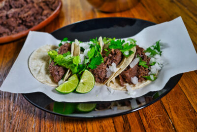 Tacos de barbacoa de lengua Cuatro tacos de barbacoa de lengua de res con cilantro, cebolla y rodajas de limón sobre tortilla, servidos en plato sobre papel.