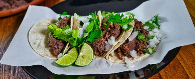 Cuatro tacos de barbacoa de lengua de res con cilantro, cebolla y rodajas de limón sobre tortilla, servidos en plato sobre papel.