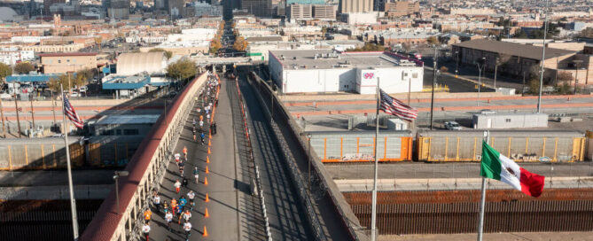 Participantes corren sobre el Puente Internacional Paso del Norte durante la carrera pedestre binacional Juárez–El Paso, con banderas de México y Estados Unidos ondeando.