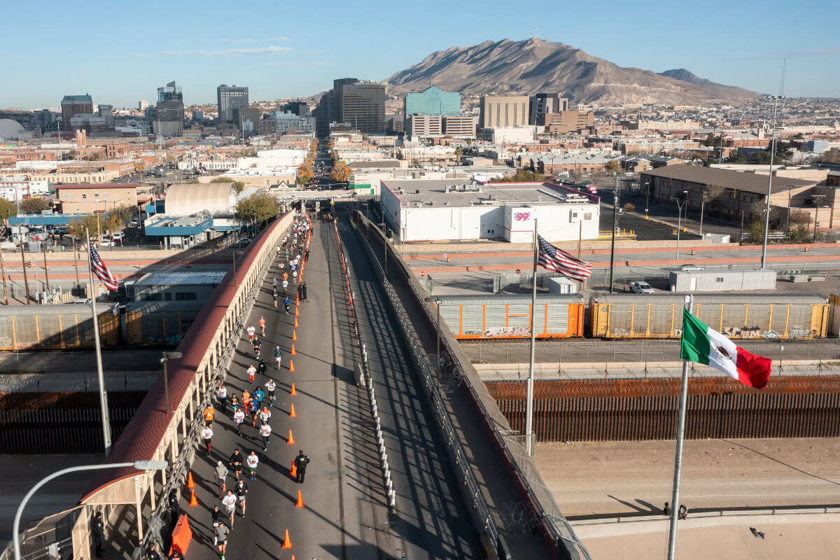 Participantes corren sobre el Puente Internacional Paso del Norte durante la carrera pedestre binacional Juárez–El Paso, con banderas de México y Estados Unidos ondeando.