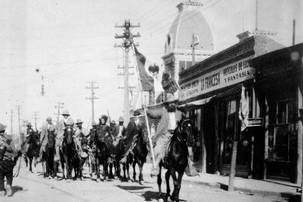 Columnas maderistas a caballo por el centro de Ciudad Juárez; a la derecha se aprecia el letrero “La Francesa” y, al fondo, la ex Aduana —hoy Museo de la Revolución en la Frontera (MUREF)—