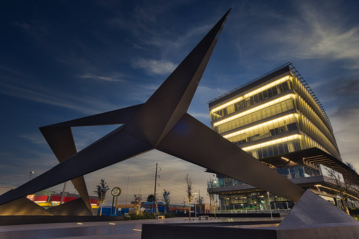 Escultura abstracta metálica en la Plaza Cantabria sobre la avenida Paseo Triunfo de la República, de noche, con edificio iluminado de fondo.