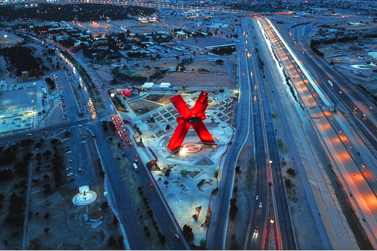Vista aérea nocturna del Monumento a la Mexicanidad, La X, en la Plaza de la Mexicanidad de Ciudad Juárez, rodeado de avenidas iluminadas.