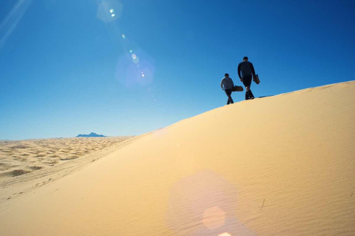 Dos personas caminando rumbo a la cima de una gran duna en el desierto — preparándose para descender en tabla de sandboard.